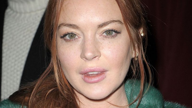 Lindsay Lohan in einem grünen Mantel, der mit neutralem Ausdruck direkt in die Kamera blickt